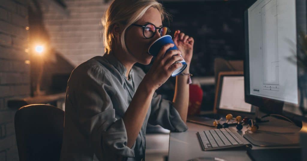 Photo of a young woman sitting at her desktop computer, having some hot coffee to help her trough a late night shift